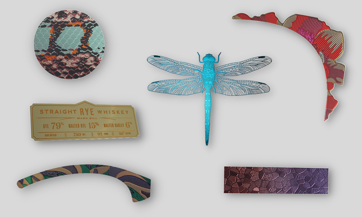 Collage of low profile electroform parts. Circular snake skin texture trim piece with sea foam green, rose gold, and brown shades. Whistle Pig whiskey label, gold with bronze copy. Arced eyebrow decorative trim piece with various textures on rose gold, green, and blue shades. Metallic blue dragonfly with microscopic openings. Faded pink to purple rectangle accent piece. Arced eyebrow corner decorative trim piece with red, purple, pink, and silver tones.